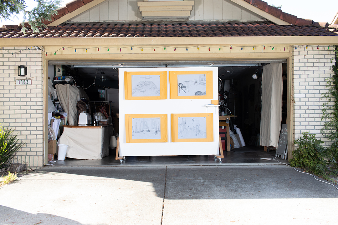 A suburban garage with an open garage door, framing a large wall with four drawings on blue paper, hung over large yellow envelopes.
