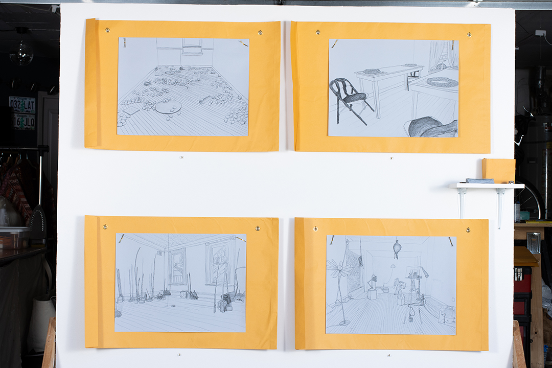 A large wall with four drawings on blue paper, hung over large yellow envelopes; a small shelf is hung on the right edge of the wall.