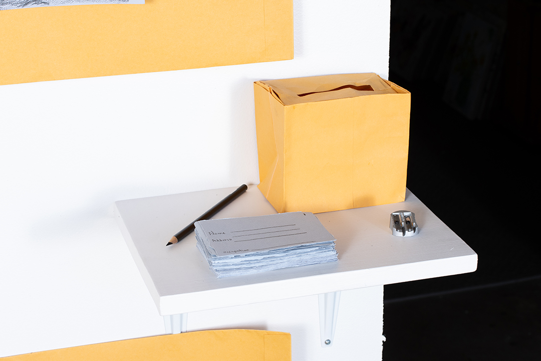 A small shelf with a box made out of yellow manila envelope paper with a slot on the top. a stack of blue paper cards with field for a name, address, and occupation, a black pencil, and a pencil sharpener.
