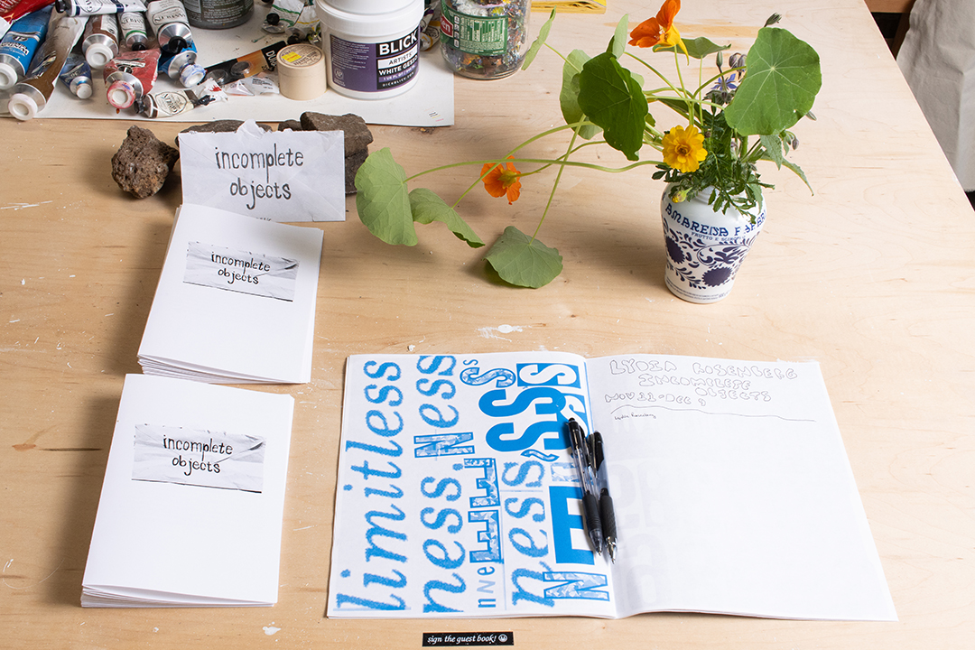 The surface of a table with a vase of flowers, pamphlets with an image of an envelope with text reading Incomplete Objects and an open paper zine.